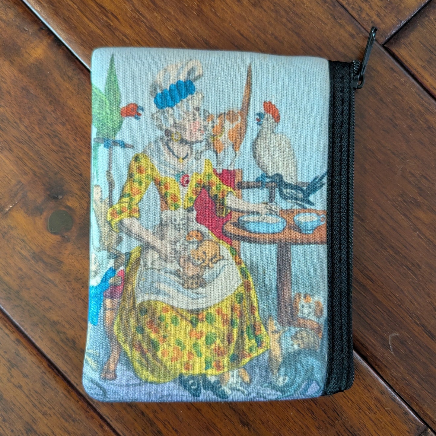 Small zipper pouch printed a woman surrounded by birds, cats, dogs, puppies and kittens while wearing a brightly colored yellow orange and green spotted dress and tall white cap with blue ribbon. From the 1790s. Virgil's Fine Goods