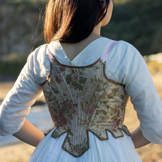Back View of gold, green and pink floral early 1800s corset (known as stays) that ends just above natural waist with pink ribbon straps. Worn by a person with long dark hair at the beach. She is wearing a white dress underneath. Scroop Patterns, Minette Stays Pattern, Circa 1785-1810, Virgil's Fine Goods