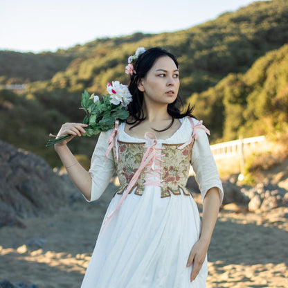 Front View of gold, green and pink floral early 1800s corset (known as stays) that ends just above natural waist with pink ribbon straps. Worn by a person with long dark hair at the beach. She is wearing a white dress underneath. Scroop Patterns, Minette Stays Pattern, Circa 1785-1810, Virgil's Fine Goods
