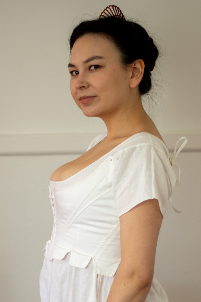 Side View Option A of white early 1800s corset (known as stays) that ends just above natural waist with pink ribbon straps. Worn by a person with long dark hair at the beach. She is wearing a white dress underneath. Scroop Patterns, Minette Stays Pattern, Circa 1785-1810, Virgil's Fine Goods