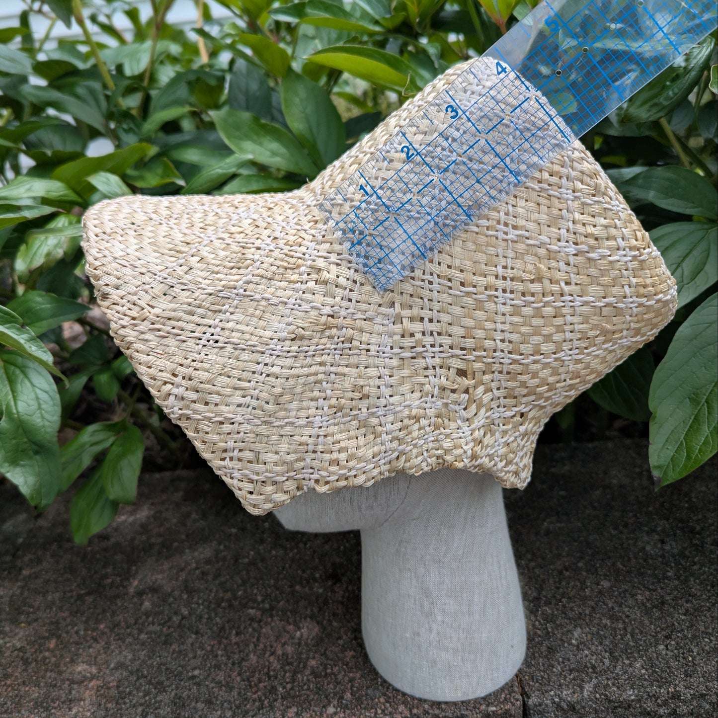 Side View Tan and white Straw bonnet with squared crown and open brim for regency era, being measured by a ruler showing a 4" high crown. Background of green peony leaves. VirgilsFineGoods.com