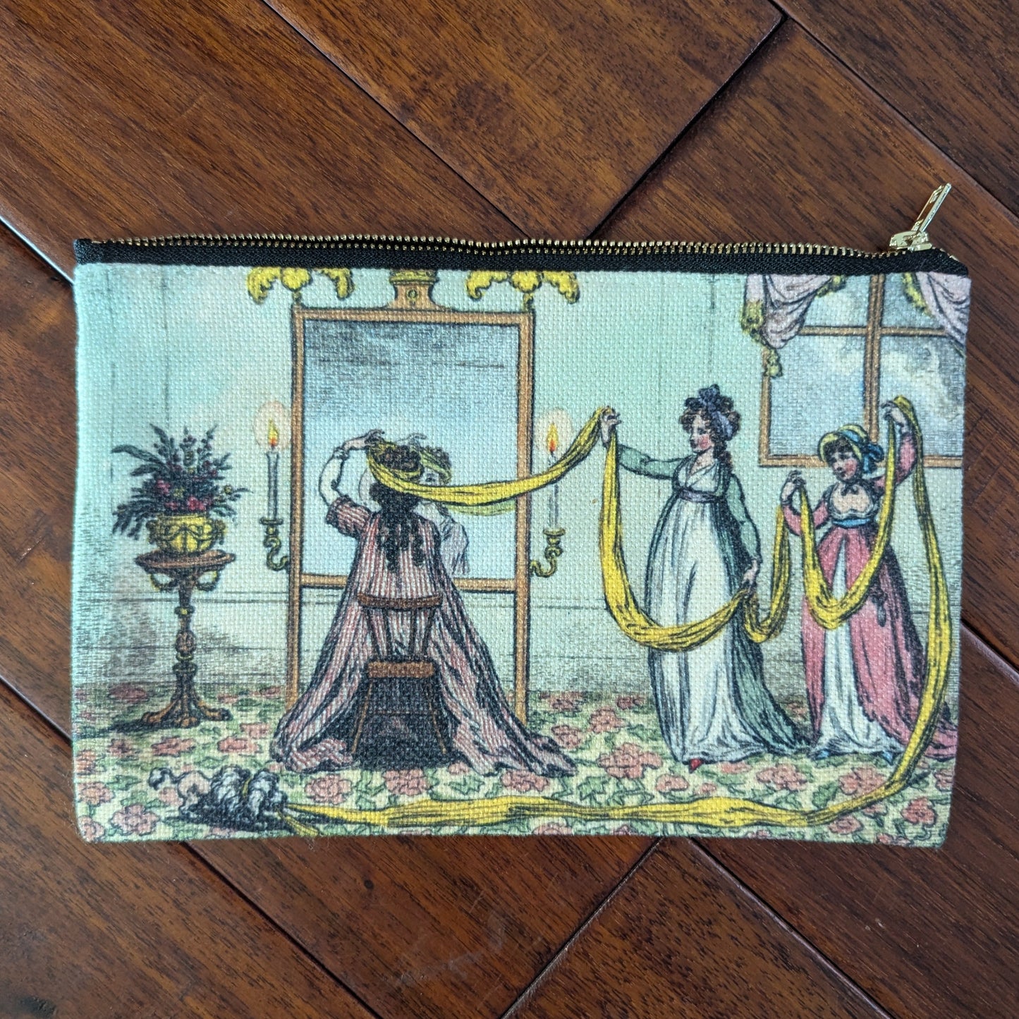 Large zipper pouch printed with 3 women with a long yellow scarf to help a woman sitting at a mirror with wrapping hair for turban. From the 19th century with bright dresses and aqua colored room with floral floor. Virgil's Fine Goods